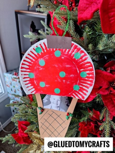 A handmade Christmas ornament, perfect for CHRISTMAS CRAFTS FOR KIDS, is shaped like an ice cream cone with a red and white top and green dots. It hangs on a decorated tree and features a small black-and-white photo of a child.