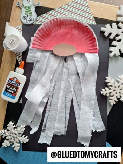 A Santa Claus craft, perfect for CHRISTMAS CRAFTS FOR KIDS, uses a red paper plate, white tissue beard strips, an oval face, and festive supplies like glue and a snowflake—all arranged neatly on a table.