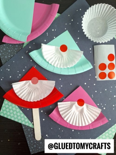 Colorful paper plate and cupcake liner fans, perfect for CHRISTMAS CRAFTS FOR KIDS, are decorated with red dot stickers and arranged on patterned paper. Craft supplies like sticks, liners, and stickers surround the festive display. Text reads @GLUEDTOMYCRAFTS.
