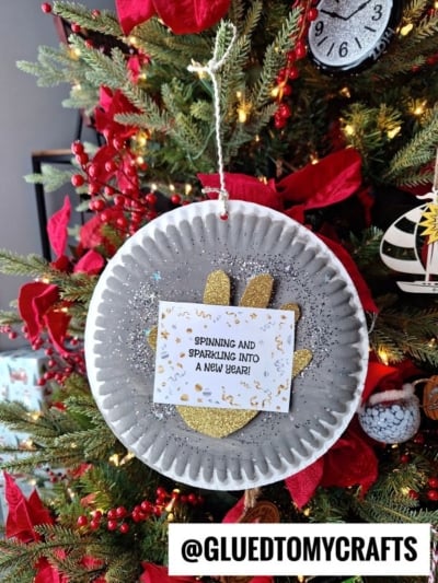 A handmade ornament hangs on a Christmas tree. It features a gray paper plate disco ball with glitter, a gold glitter paw print, and a card that says, Spinning and sparkling into a New Year! Red decorations surround it.
