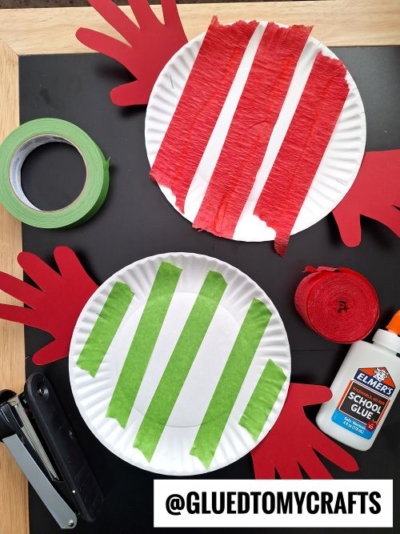 Two paper plates with handprint cutouts as handles, decorated with red and green striped crepe paper. Nearby are green tape, red crepe streamer, glue, and a stapler—perfect materials for fun Christmas crafts for kids.
