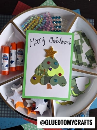 A handmade Christmas card with a tree made of green paper hearts, decorated with sequins and a gold star. Perfect for CHRISTMAS CRAFTS FOR KIDS, with glue sticks, beads, and paper cutouts arranged neatly in a circular tray underneath.