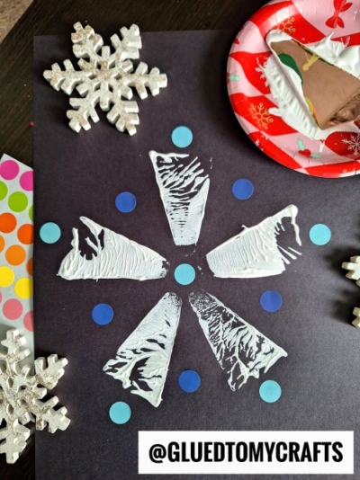 Black paper features a textured white snowflake pattern made from painted triangles, some stamped with a pie server, and is surrounded by blue dot stickers. Glittery snowflake ornaments and a plate with painted sponge are nearby. @GLUEDTOMYCRAFTS is at the bottom.