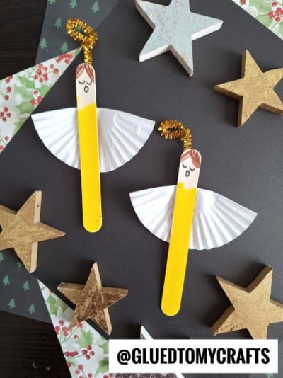 Two angel crafts made from yellow-painted popsicle sticks, white cupcake liner wings, drawn faces, and gold pipe cleaner halos are surrounded by wooden stars on a dark background—perfect CHRISTMAS CRAFTS FOR KIDS.