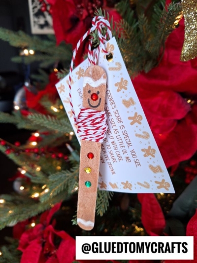 A popsicle stick gingerbread man height ornament, adorned with colored gems, twine, and a smiling face, hangs on a Christmas tree near a festive tag featuring a gingerbread cookie poem. Red poinsettias brighten the background.