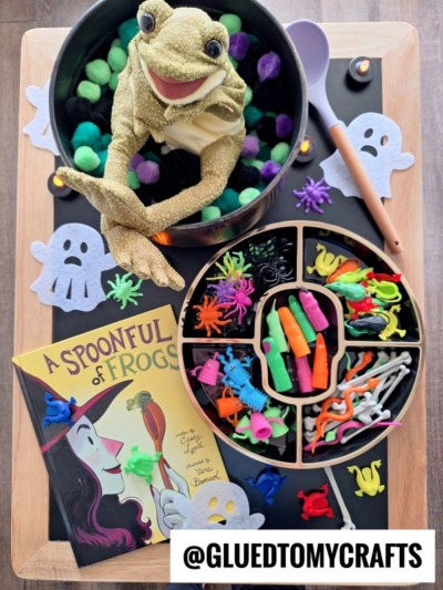 A fun Halloween craft setup inspired by fall themed sensory bin ideas, featuring a toy frog in a cauldron with green and black pom-poms, toy insects and snakes, ghost cutouts, a purple spoon, and the book A Spoonful of Frogs.