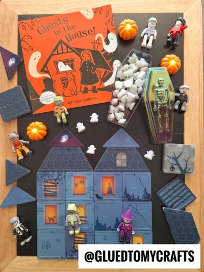 Flat lay of Halloween-themed items, including the book Ghosts in the House!, skeleton and monster figurines, ghost-shaped candies, a toy haunted house, plastic coffins, and pumpkins—perfect for FALL THEMED SENSORY BIN IDEAS—on a black background.