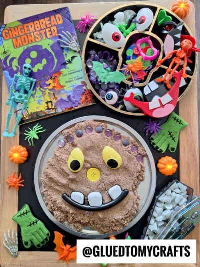 A Halloween-themed sensory activity setup inspired by FALL THEMED SENSORY BIN IDEAS featuring a Gingerbread Monster book, toy skeletons, pumpkins, monster craft supplies, and a sand tray with a monster face made from buttons and shapes.