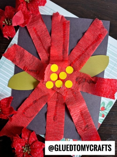 A festive craft flower made with red crepe paper strips and yellow dots on black paper, gold paper leaves, and surrounded by red silk flowers. Perfect for CHRISTMAS CRAFTS FOR KIDS, with notebook paper beneath and a @GLUEDTOMYCRAFTS watermark.