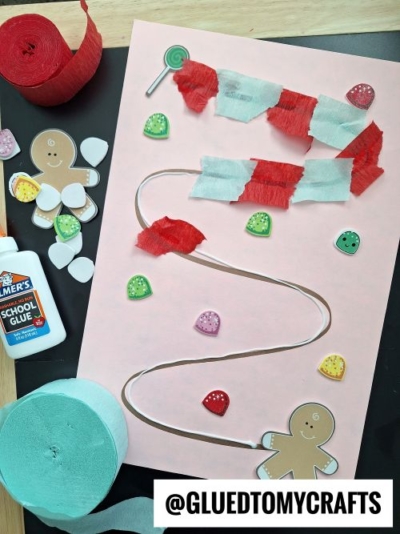 A gingerbread man craft on pink paper shows a maze path with candy and torn red-and-white paper strips as obstacles, making it a fun Christmas crafts for kids activity. Surrounded by glue, crepe paper, candies, and a gingerbread man cutout. @GLUEDTOMYCRAFTS is at the bottom.