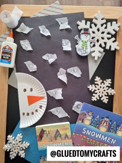 A winter-themed CHRISTMAS CRAFTS FOR KIDS setup with black paper, paper snowflakes, a snowman face made from a paper plate, glue, and children’s books Snowmen at Christmas and Snowmen at Night. @GLUETOMYCRAFTS is written at the bottom.
