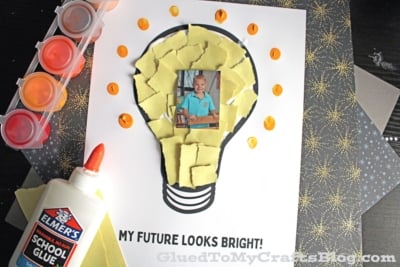 A children’s craft features a photo of a child in the center of a lightbulb outline, filled with torn yellow paper and orange dots.