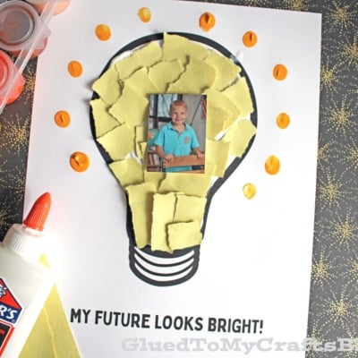 A children’s craft features a photo of a child in the center of a lightbulb outline, filled with torn yellow paper and orange dots.