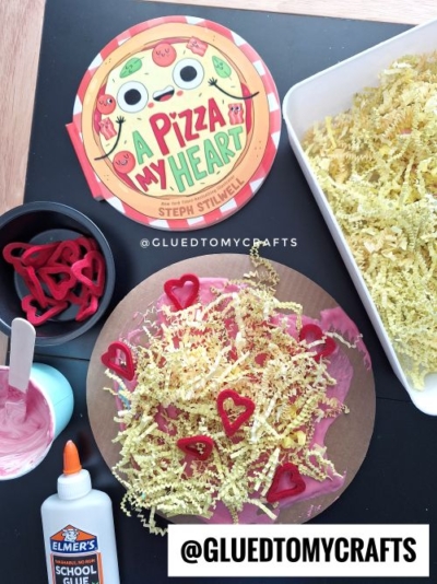 A Valentine's Day pizza craft inspired by the book "A Pizza My Heart." The craft features a circular cardboard base covered in pink puffy paint as "sauce," yellow shredded paper as "cheese," and red heart-shaped cutouts as "toppings." The setup includes a glue bottle, a bowl of pink paint, and additional heart cutouts, all displayed on a dark surface with the book nearby.