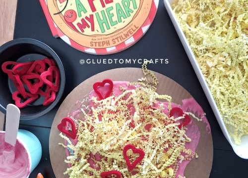 A Valentine's Day pizza craft inspired by the book "A Pizza My Heart." The craft features a circular cardboard base covered in pink puffy paint as "sauce," yellow shredded paper as "cheese," and red heart-shaped cutouts as "toppings." The setup includes a glue bottle, a bowl of pink paint, and additional heart cutouts, all displayed on a dark surface with the book nearby.