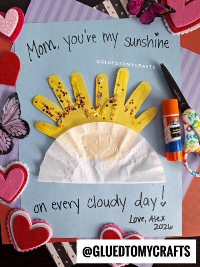 A handmade card featuring a yellow handprint sun with glitter rays and a coffee filter cloud on a light blue background. The message reads, "Mom, you're my sunshine on every cloudy day! Love, Alex 2026." The card is surrounded by felt hearts, butterflies, and craft supplies like scissors and glue. The watermark "@GLUEDTOMYCRAFTS" is visible.