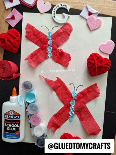 A craft project featuring two red tissue paper butterflies with blue painted bodies on white paper—perfect for valentine's day craft ideas for kids. School glue, paint, felt hearts, yarn hearts, and paper hearts are arranged around the artwork.