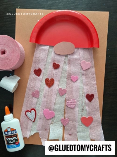 A Valentine’s Day jellyfish craft—a fun addition to Valentine’s Day craft ideas for kids—made from a red paper plate, pink paper strips, and heart-shaped decorations, with glue, pink crepe paper, and a paper base on a dark surface.