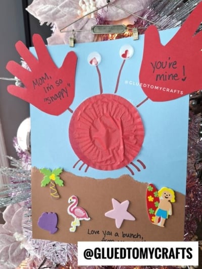 A handmade card featuring a red crab made from a cupcake liner body and red handprint claws on a beach-themed background. The message reads, "MOM, I'm so 'snappy' you're mine! Love you a bunch, from ..." The card includes foam stickers of a palm tree, seashell, flamingo, and starfish, with the watermark "@GLUEDTOMYCRAFTS.