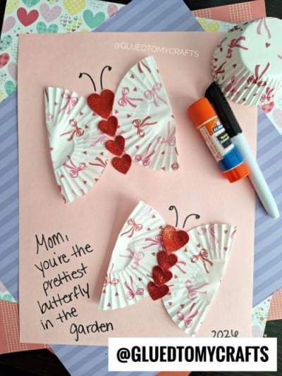 A handmade card with cupcake liner butterflies and red glitter hearts—perfect for valentine's day craft ideas for kids. Text reads, Mom, you're the prettiest butterfly in the garden. A glue stick and black marker are on the card.