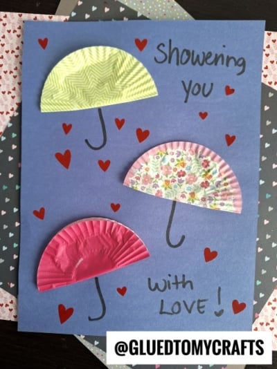 A blue paper card with three half-cut cupcake liners as umbrellas, red hearts, and "Showering you with love!" handwritten in black—@GLUEDTOMYCRAFTS at the bottom. Perfect for valentine's day craft ideas for kids.