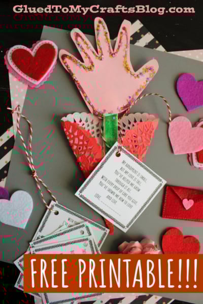Flat lay of a DIY handprint flower craft featuring a pink glittery handprint on a green craft stick as the flower stem, surrounded by decorative red and white doilies. A large white tag with a heartfelt poem about love and growth hangs from the craft, tied with red and white baker's twine. Smaller tags with similar designs are stacked beneath. The background includes gray paper, patterned papers, and scattered felt hearts in red, pink, purple, and white. A red felt envelope and pink felt rosettes add to the cheerful, sentimental aesthetic. The image promotes a free printable from GluedToMyCraftsBlog.com, perfect for creating keepsakes or gifts for occasions like Mother’s Day or Valentine’s Day.