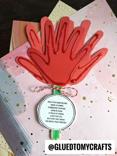 A handmade handprint flower craft made from layered red cutouts on a green stem, with a round tag featuring a handprint flower poem, placed on decorative pink paper. The handle reads @GLUEDTOMYCRAFTS.