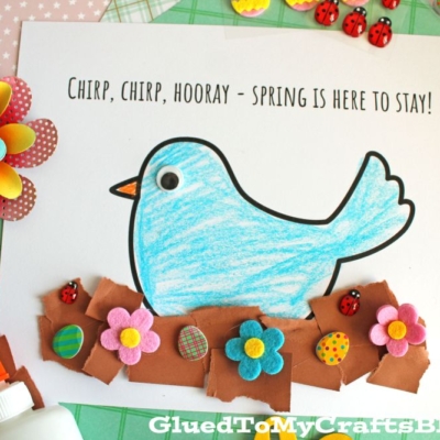 A blue paper bird with a googly eye is colored and glued on a white sheet that says, Chirp, chirp, hooray—Spring is here to stay! with colorful foam flowers and ladybugs along the brown paper ground.