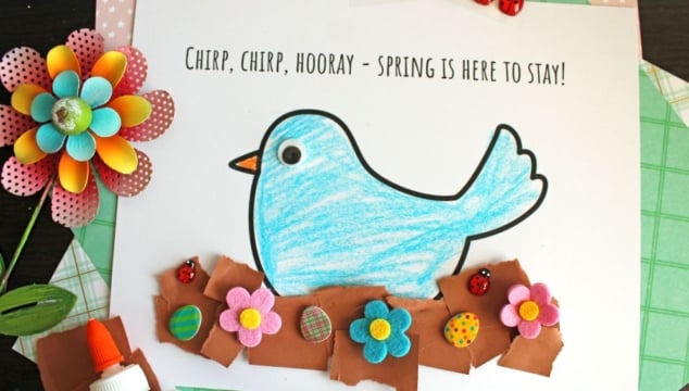 A blue paper bird with a googly eye is colored and glued on a white sheet that says, Chirp, chirp, hooray—Spring is here to stay! with colorful foam flowers and ladybugs along the brown paper ground.