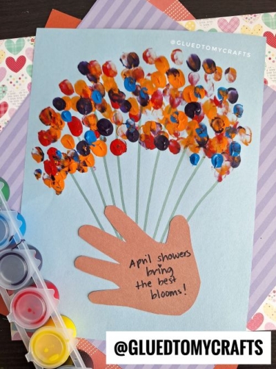A handmade card, perfect for valentine's day craft ideas for kids, features a brown paper cutout of a hand holding painted dot flowers. The text on the hand reads, "April showers bring the best blooms!" A paint palette and patterned paper complete the scene.