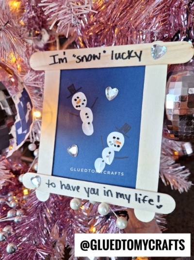 A handmade snowman craft ornament hangs on a pink Christmas tree. The frame reads I’m snow lucky to have you in my life! with two snowmen and shiny heart stickers—perfect for Valentine's Day craft ideas for kids. @GLUEDTOMYCRAFTS is at the bottom.