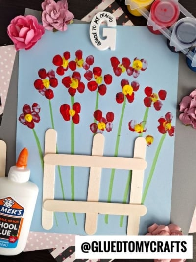 A vibrant craft featuring finger-painted red and yellow flowers with green stems on a light blue background. A small popsicle stick fence is placed at the bottom, and the craft is surrounded by paint pots, a glue bottle, artificial flowers, and decorative papers. Branding from "Glued To My Crafts" is visible.