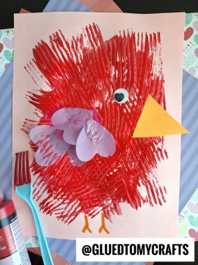 A child’s art project of a red bird made with fork-painted strokes, purple paper wings, a yellow paper beak, a googly eye, and orange legs on peach paper is a perfect addition to valentine's day craft ideas for kids, with colorful supplies nearby.