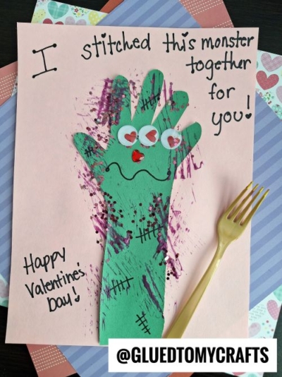 A handmade Valentine’s card features a green paper hand decorated like a monster, with googly eyes and heart stickers—one of the cutest Valentine’s Day craft ideas for kids. Black stitches, a yellow plastic fork, and painted smudges add creative flair.