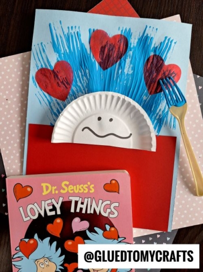 A DIY Dr. Seuss-inspired project and one of the cutest valentine’s day craft ideas for kids—using a paper plate, red hearts, blue paint strokes, a gold fork, and Lovey Things book on a polka-dot surface with a smiling face and blue hair.