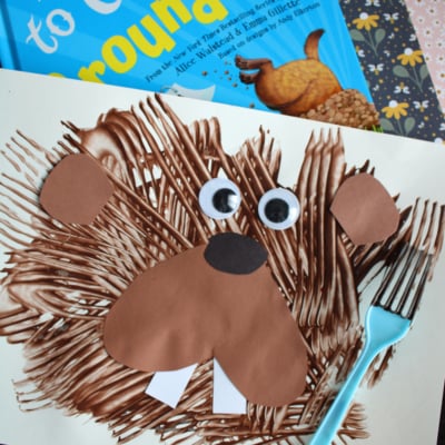 A childrens craft project features a fork painted groundhog made from brown lines on paper, with added paper ears, eyes, nose, and teeth. A blue fork and the book How to Catch a Groundhog appear in the background.