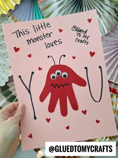 A whimsical Valentine's Day card on a pink background with a red handprint monster featuring googly eyes, antennae, and a toothy smile. The text says, "This little monster loves YOU," with red hearts scattered around. The card is held by a hand, with decorative paper fans in the background and branding from "Glued To My Crafts."