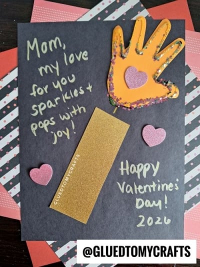 A dark gray card with an orange glitter-outlined handprint and pink glitter heart, surrounded by handwritten messages like "Mom, my love for you sparkles + pops with joy!" and "Happy Valentine's Day! 2026." The card is layered on patterned papers with pink polka dots and stripes, and the watermark "@GLUEDTOMYCRAFTS" is visible.