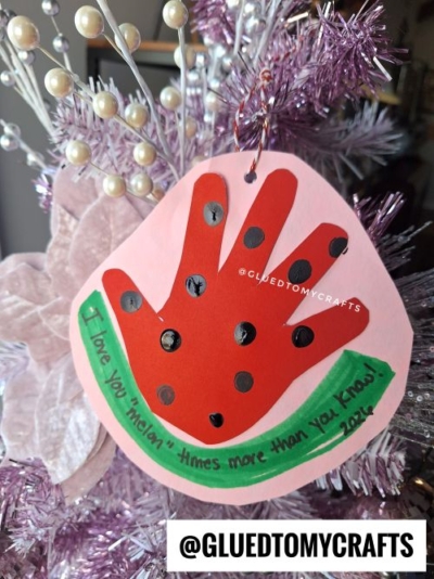 A pink ornament shaped like a watermelon slice with a red handprint and black dots hangs on a tree—perfect for Valentine's Day craft ideas for kids. Green text reads, I love you melon times more than you know! 2022. @GLUEDTOMYCRAFTS is written below.