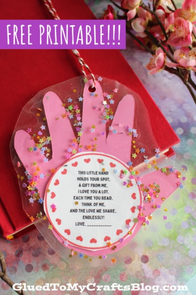 A pink handprint craft with a heart-shaped poem in the center, decorated with confetti and a twine loop, lies on a red book next to pink flowers. Text reads: FREE PRINTABLE!!! GluedToMyCraftsBlog.com.