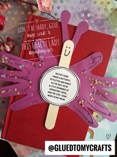 A handmade handprint popsicle stick butterfly craft with pink and purple cardstock wings, star confetti, and a smiley face. A butterfly poem is attached. Image credits @gluedtomycrafts.