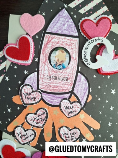 A handmade handprint rocket keepsake features a child’s photo in the window, colorful felt and paper hearts, and “I LOVE YOU BECAUSE,” with hearts listing reasons. @GLUEDTOMYCRAFTS is written at the bottom.