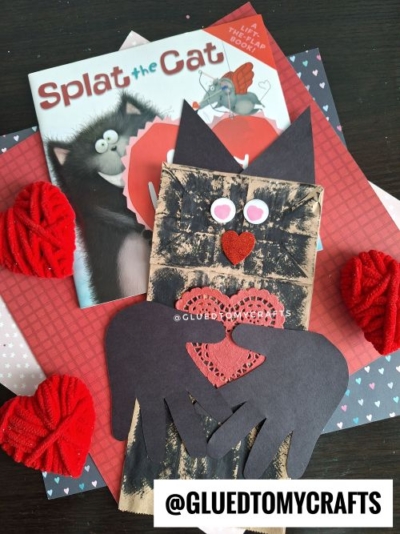 A handmade black cat craft made from a paper bag and heart-shaped cutouts is displayed on layered pink and red papers—one of the cutest Valentine's Day craft ideas for kids, with Splat the Cat and red yarn hearts in the background.