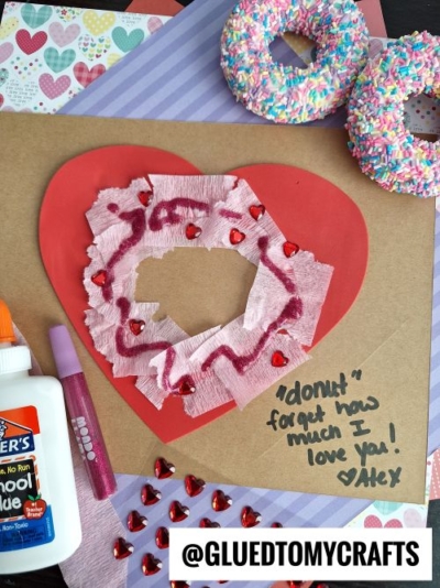 A handmade Valentines card with a red heart, tissue paper, painted swirls, red heart stickers, and the message: “‘donut’ forget how much I love you! ♥ Alex.” Perfect inspiration for Valentine's Day craft ideas for kids—craft supplies and donuts nearby.