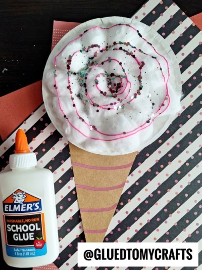 A candy-themed craft featuring a fluffy "cotton candy" design made from a coffee filter filled with cotton balls, decorated with pink squiggles and glitter. The cone is a brown paper triangle with pink stripes. The craft is displayed on a patterned background with a glue bottle nearby.