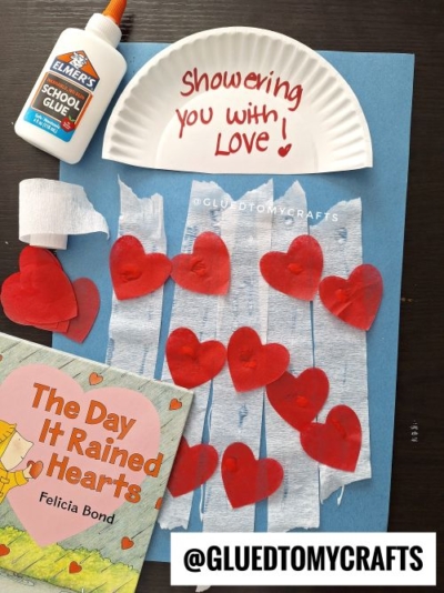 A kids’ Valentine’s Day craft features red paper hearts on blue paper with white streamers, a plate reading “Showering you with love,” glue, and “The Day It Rained Hearts.” @GLUEDTOMYCRAFTS shares perfect Valentine’s Day craft ideas for kids.