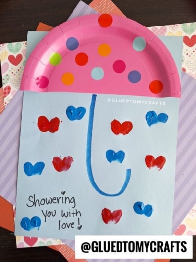 A craft shows a pink paper plate umbrella on blue paper with blue and red heart-shaped raindrops and “Showering you with love!” written in marker—a sweet option for Valentine's Day craft ideas for kids. Image watermarked @GLUEDTOMYCRAFTS.