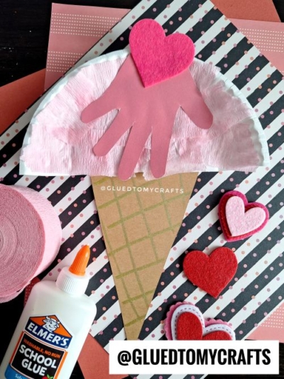 A Valentine's Day craft resembling an ice cream cone, made with a brown paper triangle cone, a pink crinkled tissue paper scoop, a pink handprint, and a bright pink felt heart on top. Surrounding the craft are a glue bottle, pink crepe paper, and felt hearts, all set on a patterned background with black and white stripes and pink dots.