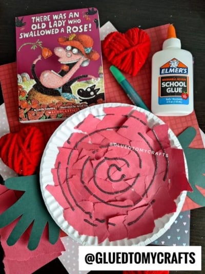 A children’s craft project is displayed with a paper plate rose made from torn pink paper, a bottle of Elmer’s glue, a green marker, red yarn hearts, and the book There Was an Old Lady Who Swallowed a Rose!—a perfect Valentine’s Day craft idea for kids.