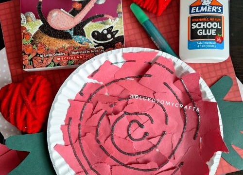 A children’s craft project is displayed with a paper plate rose made from torn pink paper, a bottle of Elmer’s glue, a green marker, red yarn hearts, and the book There Was an Old Lady Who Swallowed a Rose!—a perfect Valentine’s Day craft idea for kids.